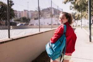 Joven estudiante sonriendo mientras camina hacia la escuela, llevando una mochila azul y vistiendo una chaqueta roja.