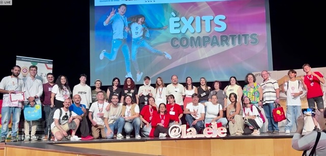 Grupo de jóvenes con camisetas blancas y rojas posando sonrientes en el escenario de Éxitos Compartidos Dénia, con el cartel del evento de fondo y el logo luminoso de La EJE.