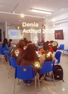 Grupo de jóvenes sentados en círculo en un aula moderna, con una proyección en la pared y símbolos de ideas y cohetes flotando. El texto "Denia Actitud 2025" aparece sobre la escena.